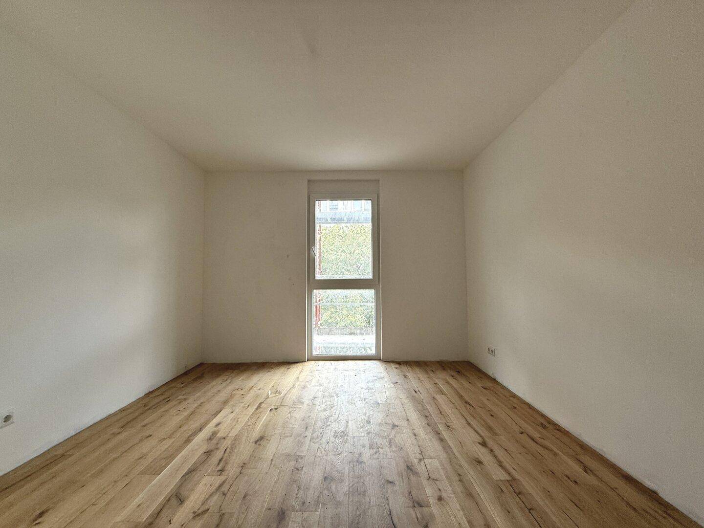 Leeres Schlafzimmer mit hellem Holzboden und einem hohen Fenster, das viel Tageslicht hereinlässt.