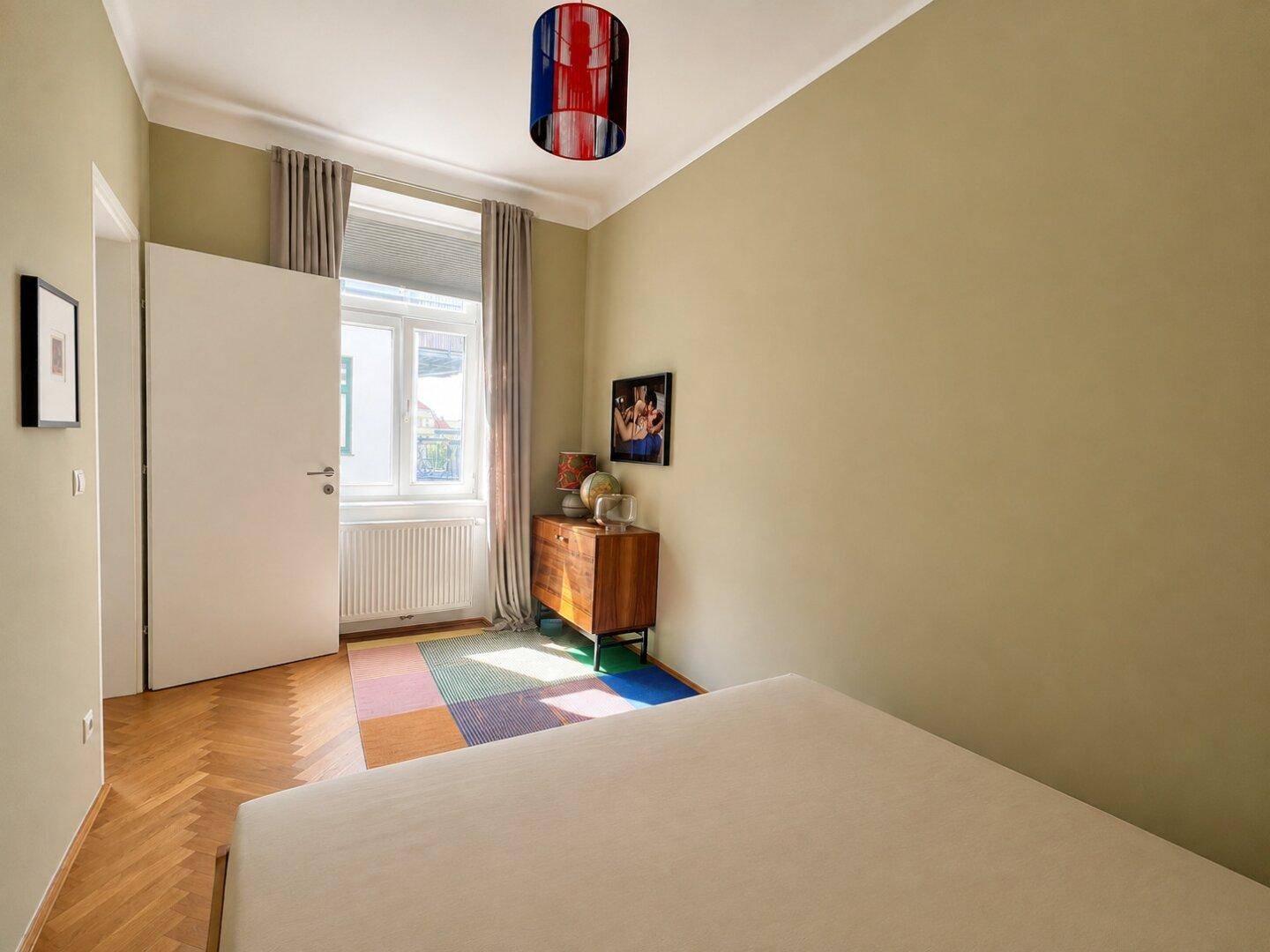 Helles Schlafzimmer mit Fischgrätparkett, beigen Wänden und farbigem Teppich.