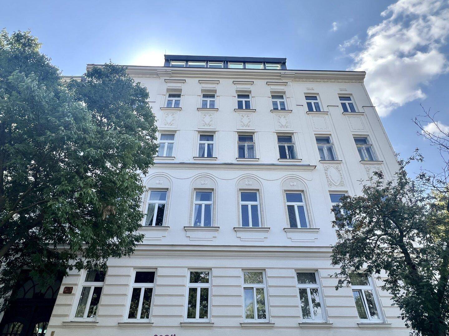Klassische Fassade eines Altbaus mit vielen Fenstern und grünen Bäumen.