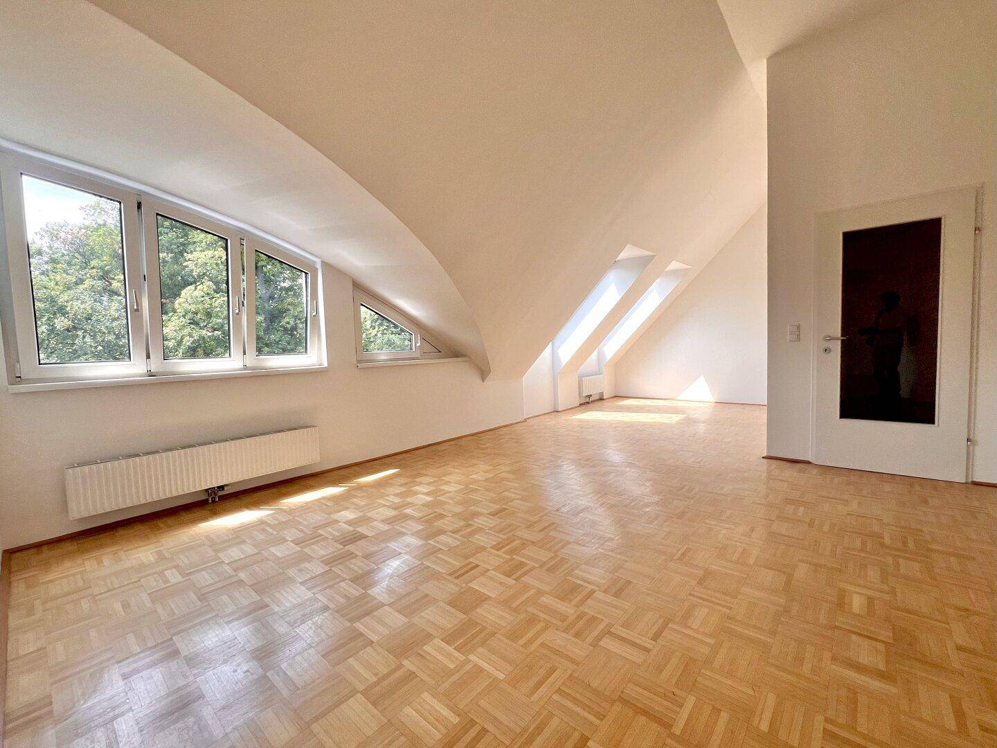 Lichtdurchfluteter Raum im Dachgeschoss mit Parkettboden, Dachschrägen und mehreren Fenstern.