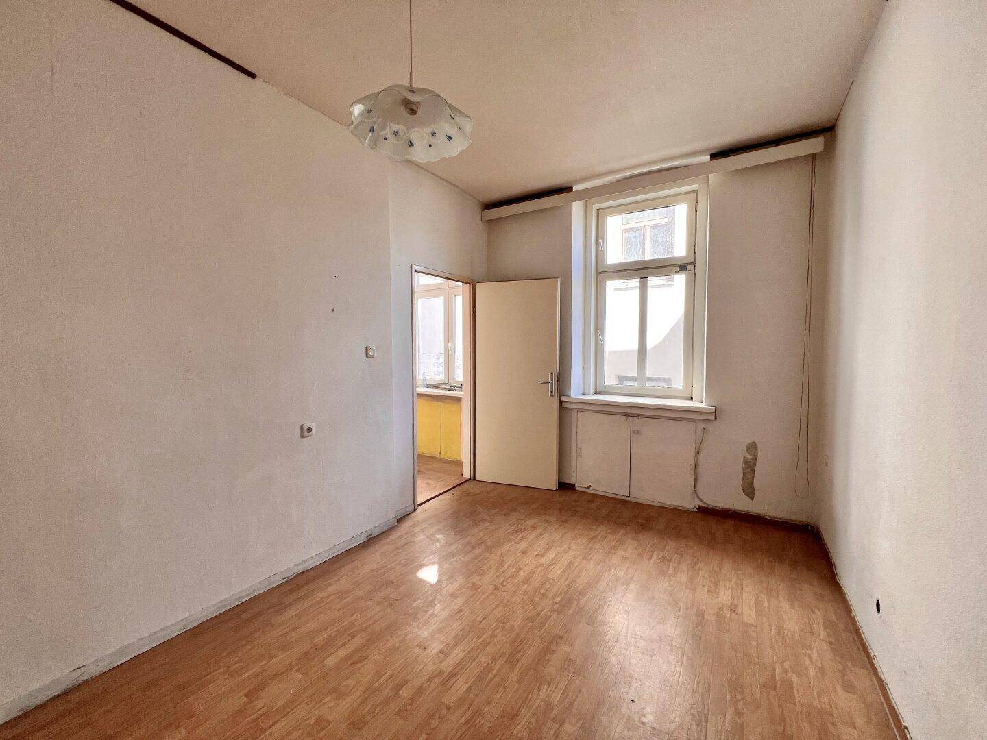 Helles, leeres Zimmer mit Holzboden und einem Fenster, das Tageslicht hereinlässt.