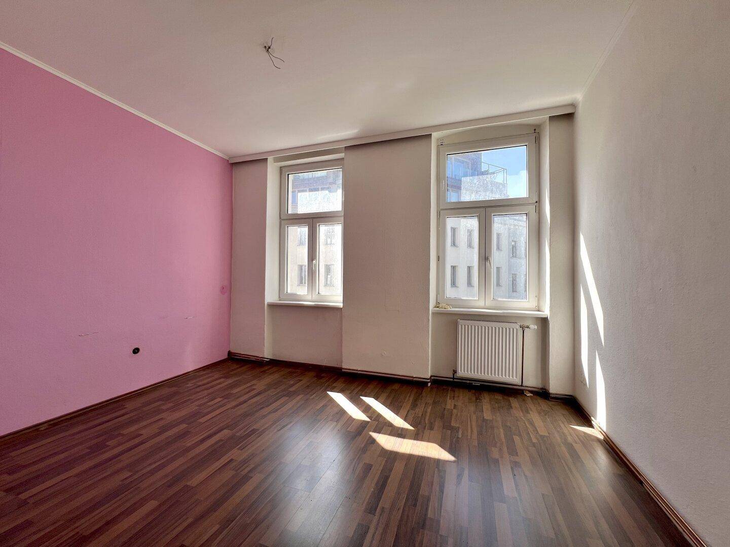Geräumiges Zimmer mit dunklem Holzboden und einer rosa Akzentwand, hell durch Tageslicht.