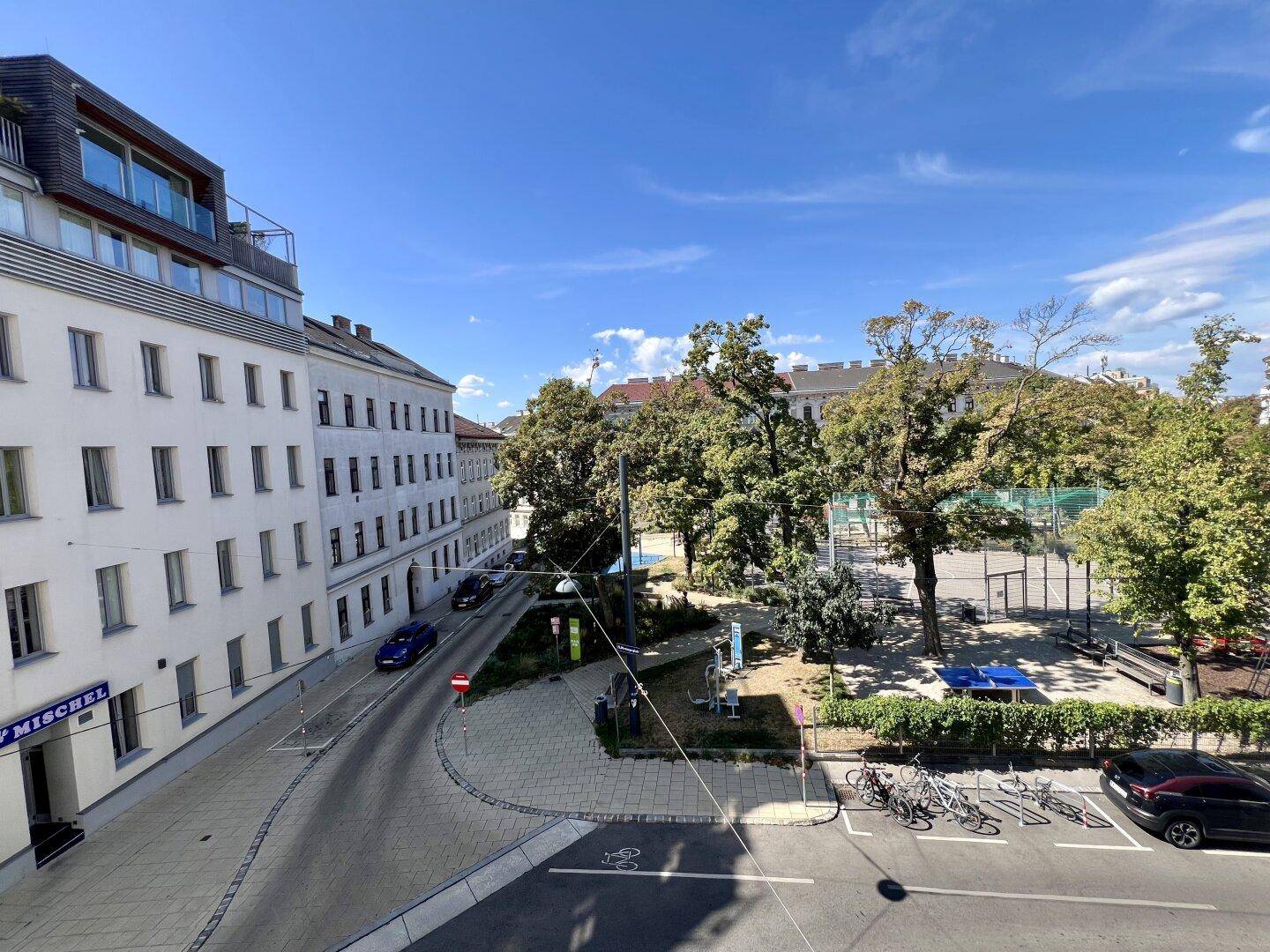 Blick auf eine belebte Straße mit angrenzendem Park und umliegenden Wohngebäuden unter blauem Himmel.