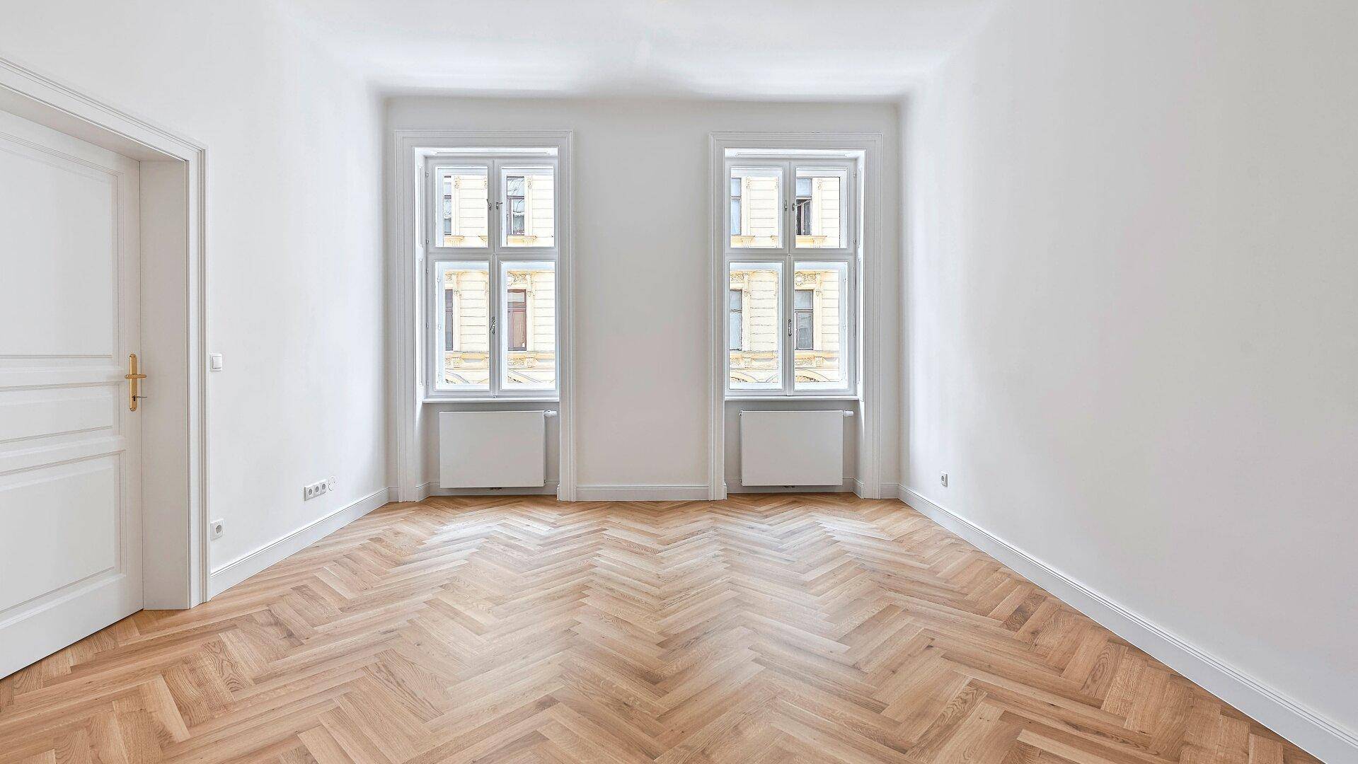 Helles Zimmer mit Fischgrätparkettboden und zwei großen Fenstern, ideal für Wohn- oder Schlafbereich.