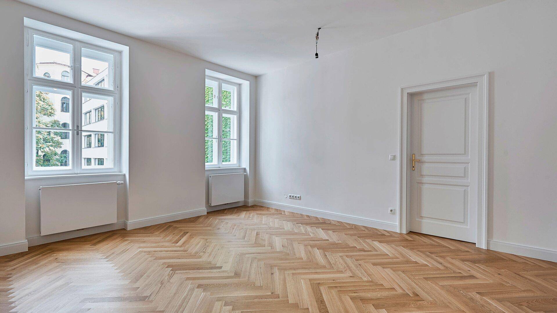 Geräumiges Zimmer mit hellem Fischgrätparkett und Fenstern an zwei Seiten für viel Tageslicht.