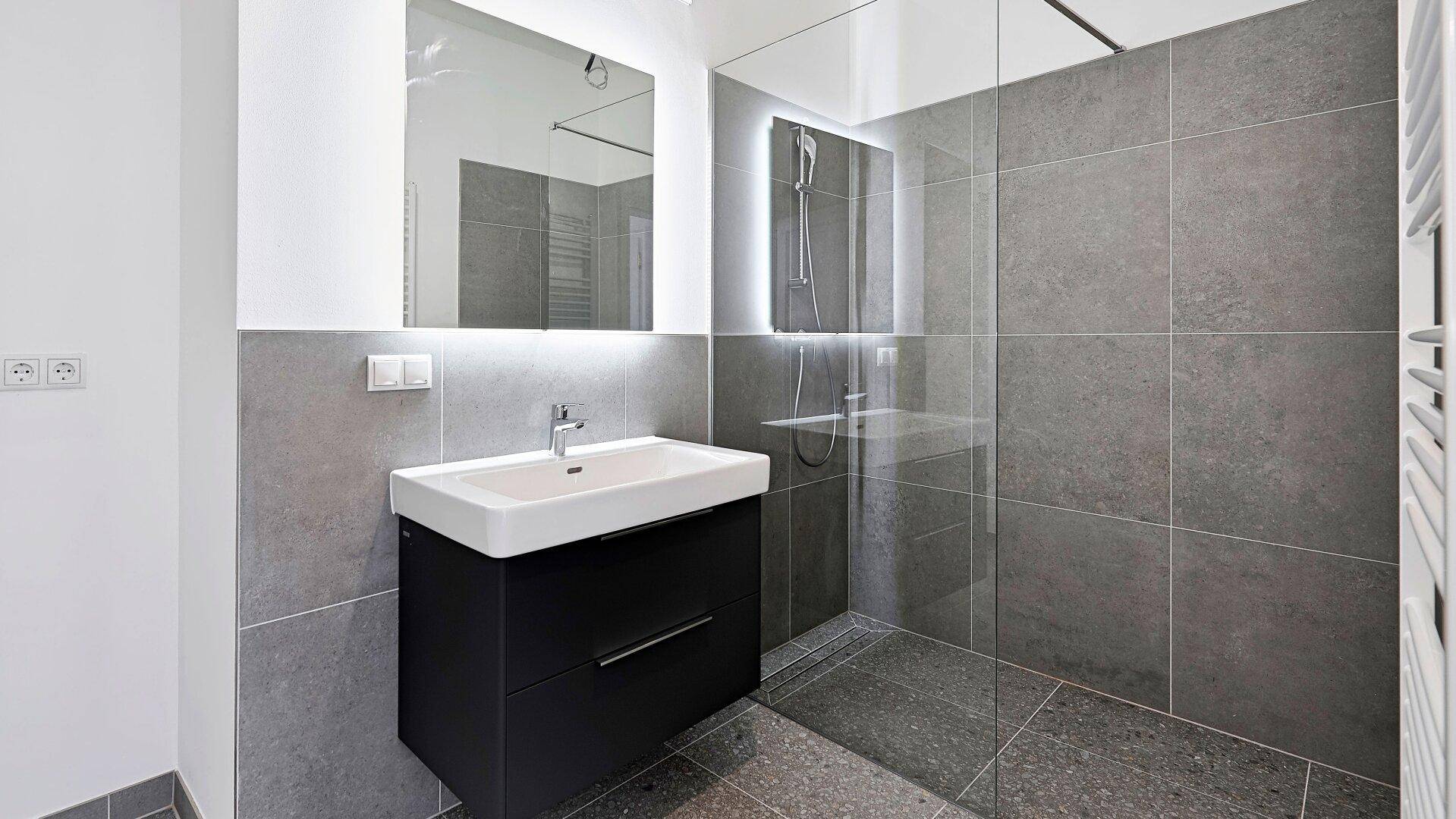 Modernes Badezimmer mit grauen Fliesen, Waschtisch, beleuchtetem Spiegel und einer geräumigen begehbaren Dusche.