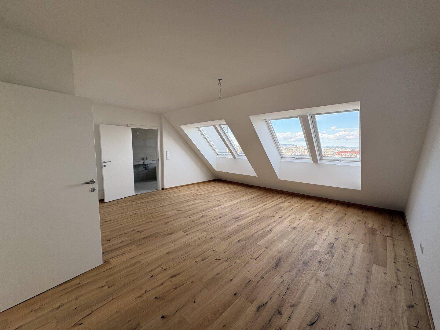 Helles Schlafzimmer im Dachgeschoss mit Holzboden, Dachfenstern und Zugang zum Badezimmer.