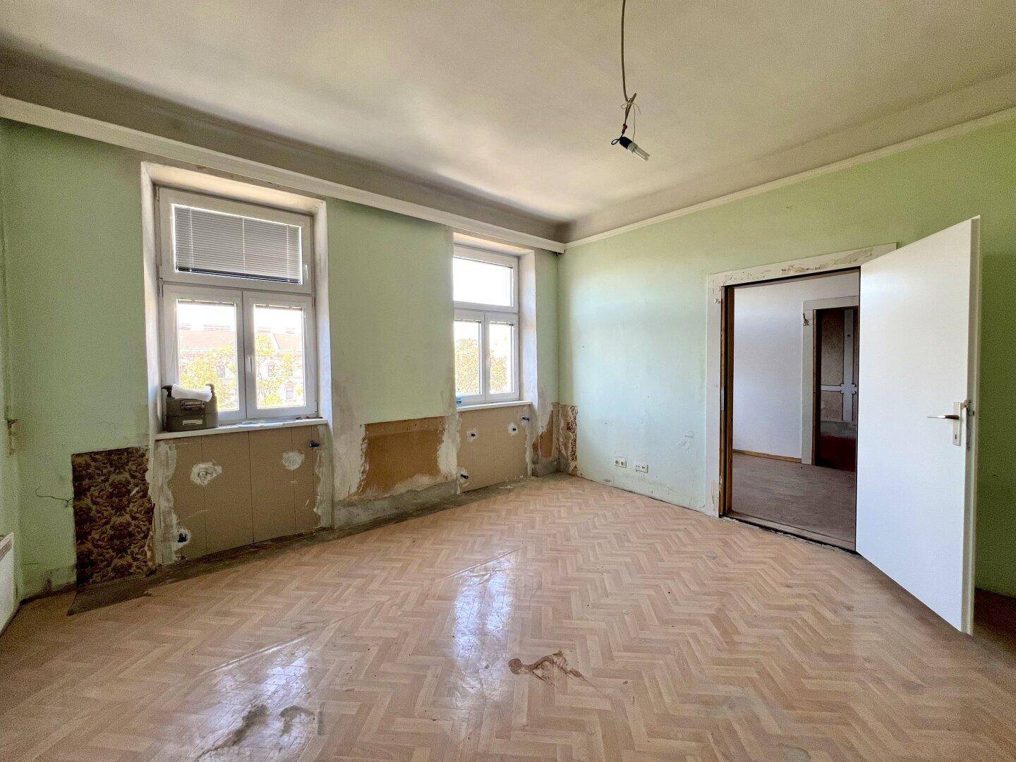 Renovierungsbedürftiger Raum mit Fischgrätparkett, hellgrünen Wänden und zwei Fenstern.