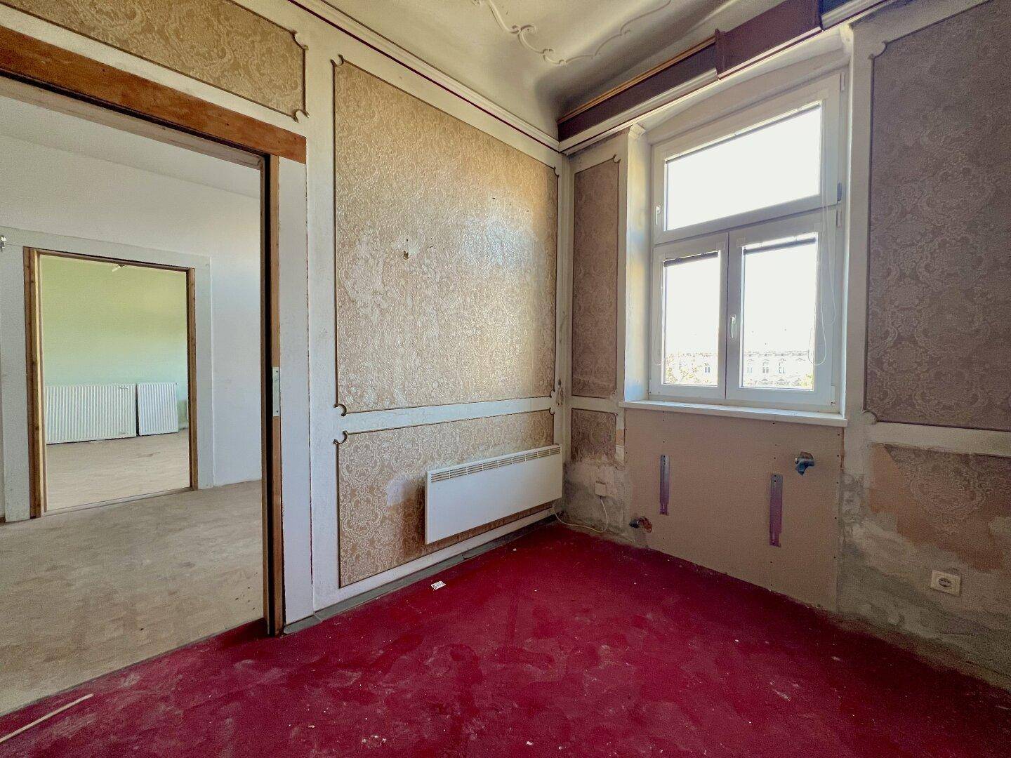 Renovierungsbedürftiger Raum mit rotem Teppichboden, gemusterter Tapete und Heizkörper.