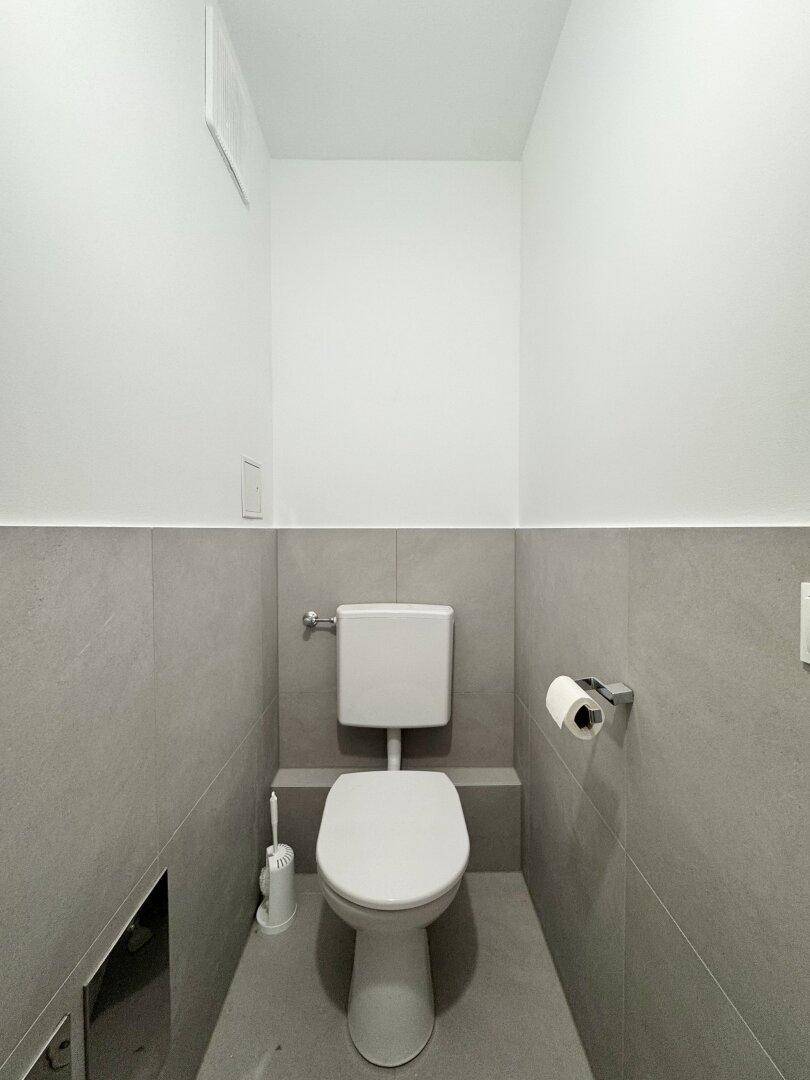 Separates WC mit wandhängender Toilette und modernen grauen Fliesen im unteren Bereich.