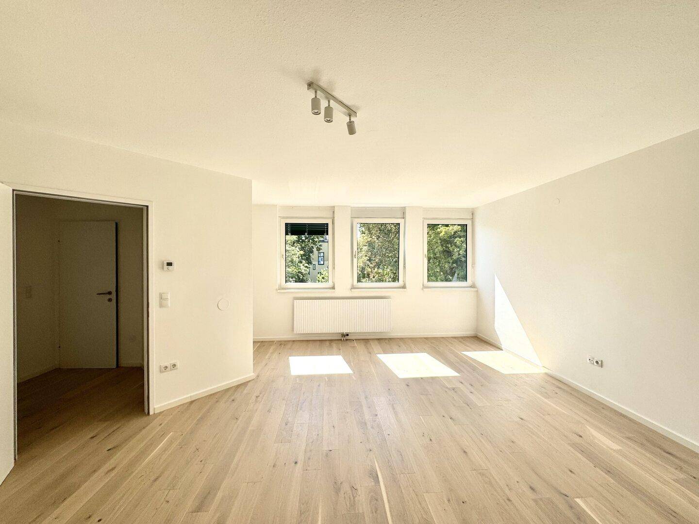 Geräumiges, helles Zimmer mit hellem Holzfußboden und Blick auf grüne Bäume.
