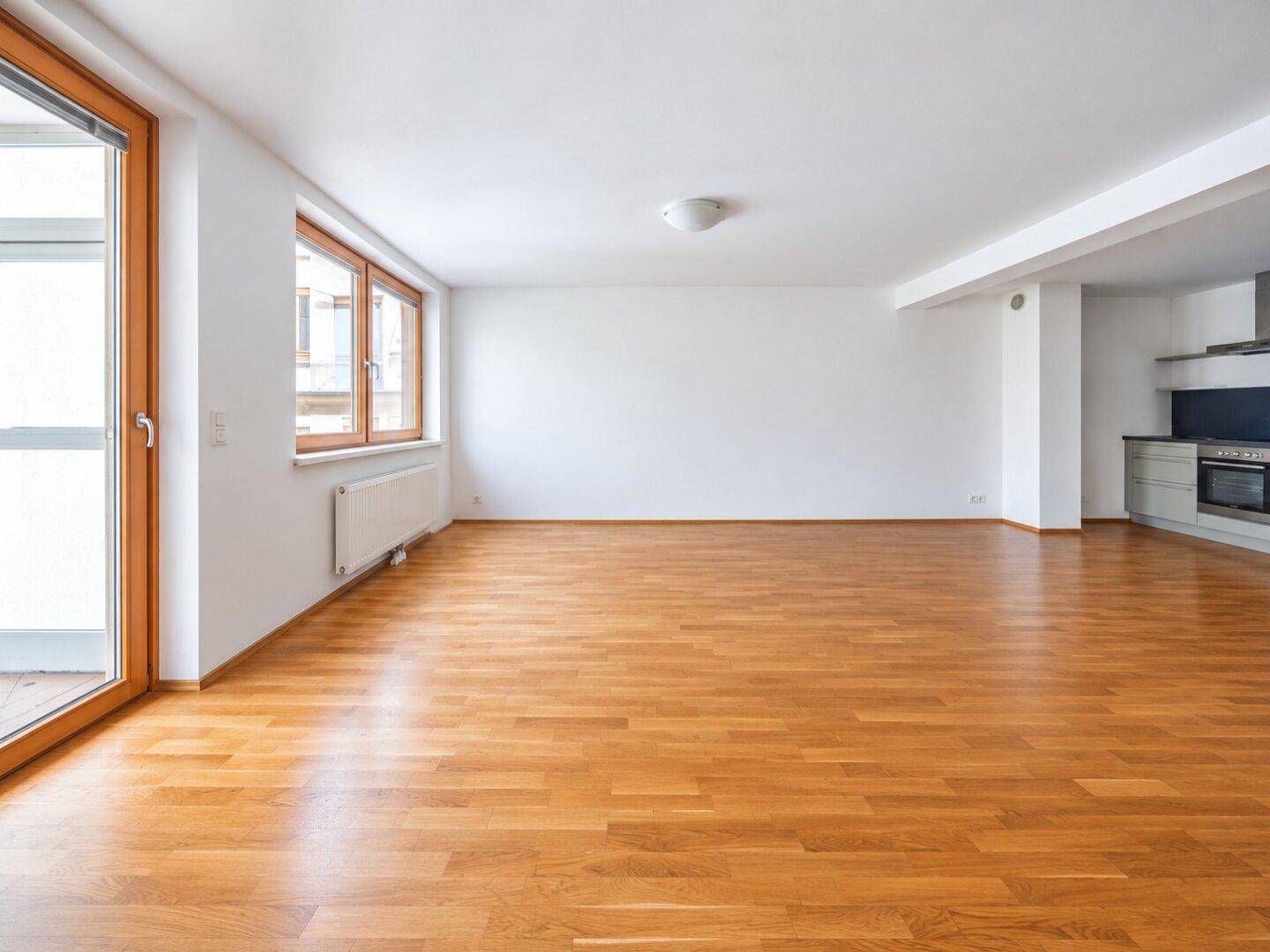 Heller, leerer Wohnbereich mit großen Fenstern, Holzparkett und Zugang zu einem Balkon.