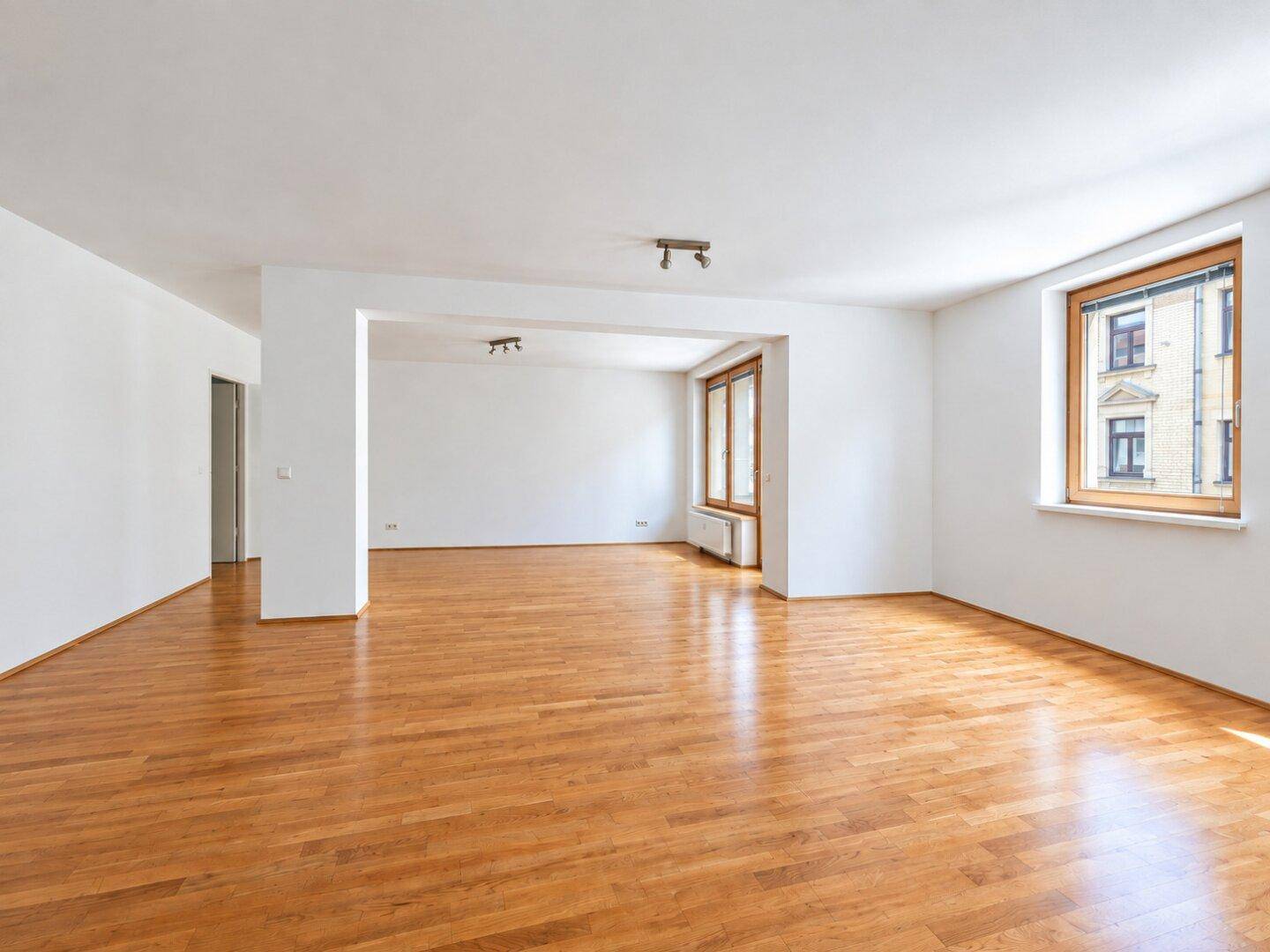 Heller Wohnraum mit Holzparkett und großen Fenstern, der eine offene und einladende Atmosphäre schafft.