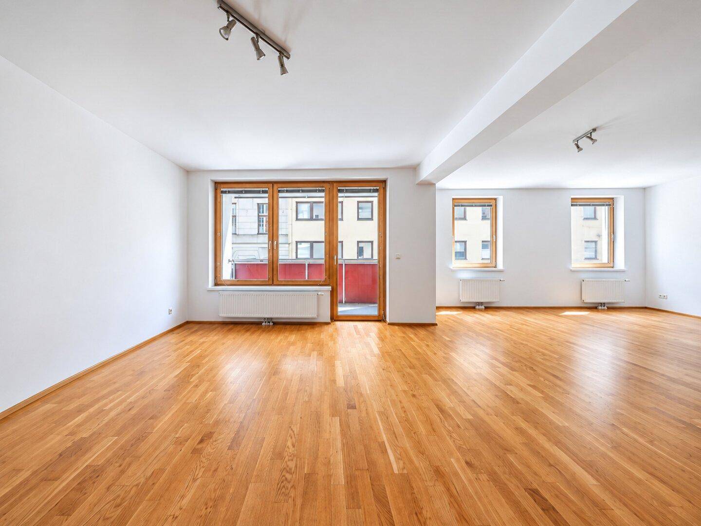 Großer, heller Wohnbereich mit Holzparkett und Fensterfront, die Zugang zum Balkon bietet.