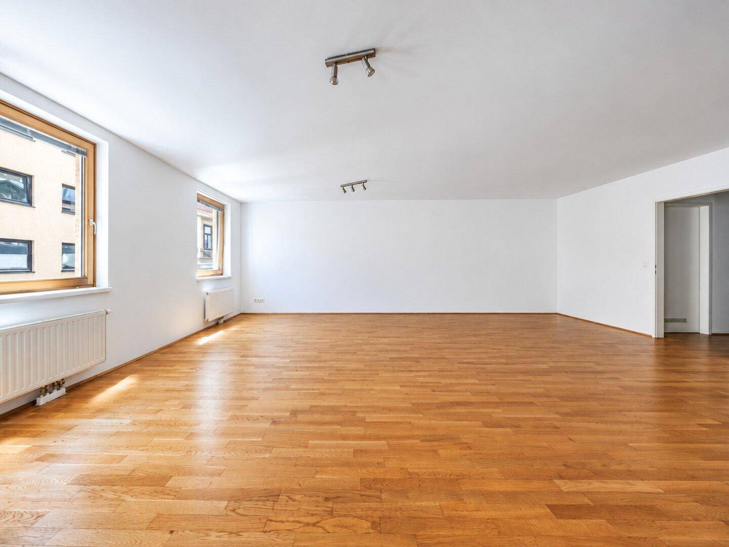 Geräumiger, heller Wohnbereich mit Holzparkett und mehreren Fenstern, die viel Tageslicht hereinlassen.