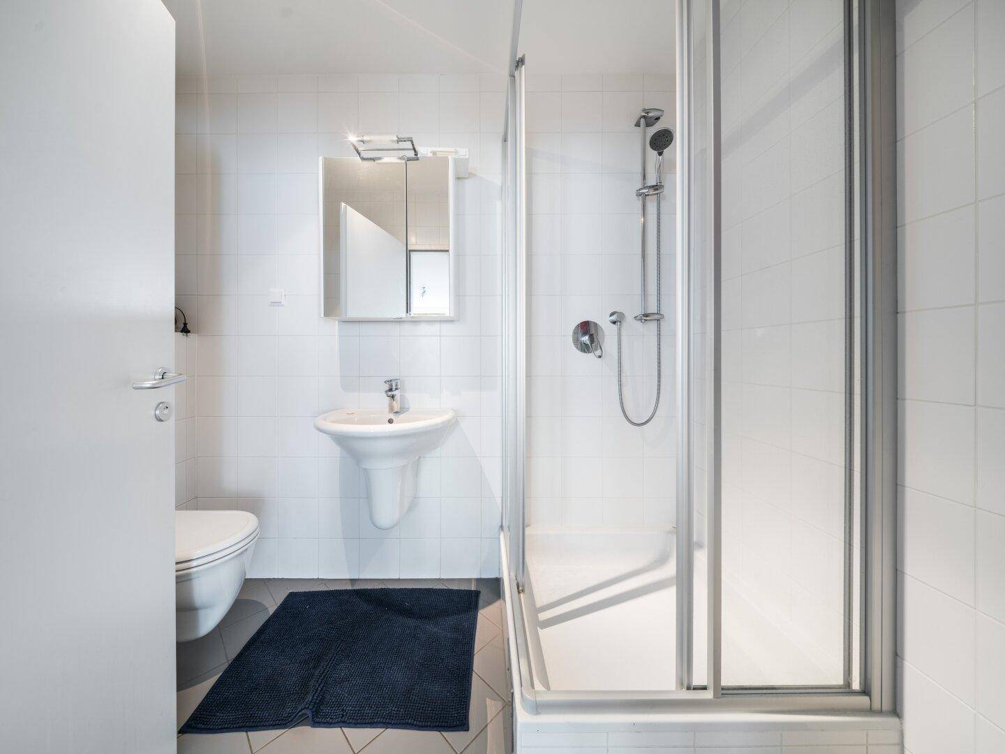 Modernes Badezimmer mit weißen Fliesen, Dusche, WC und Waschbecken mit Spiegelschrank.
