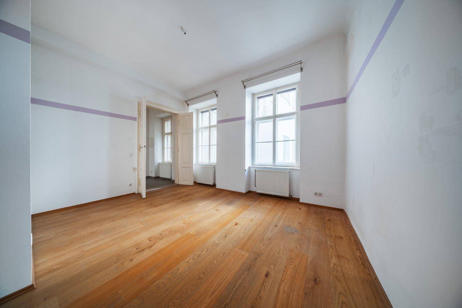 Heller, leerer Raum mit Holzdielenboden, hohen Decken und großen Fenstern, ideal als Wohnzimmer.