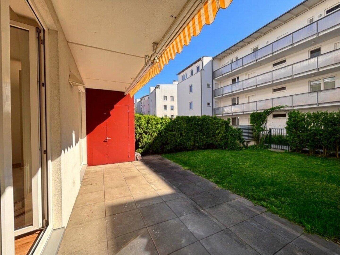 Sonniger Terrassenbereich mit Fliesenboden und angrenzender Rasenfläche, umgeben von Hecken.
