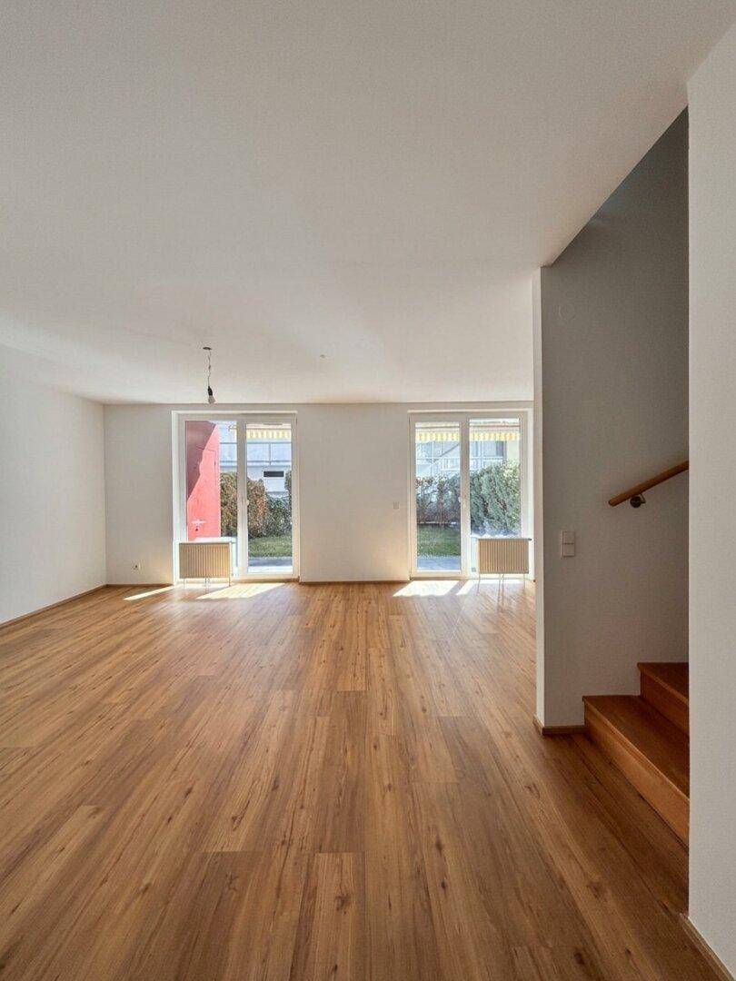 Heller, leerer Wohnbereich mit Holzboden und Treppenaufgang, mit Blick ins Grüne.