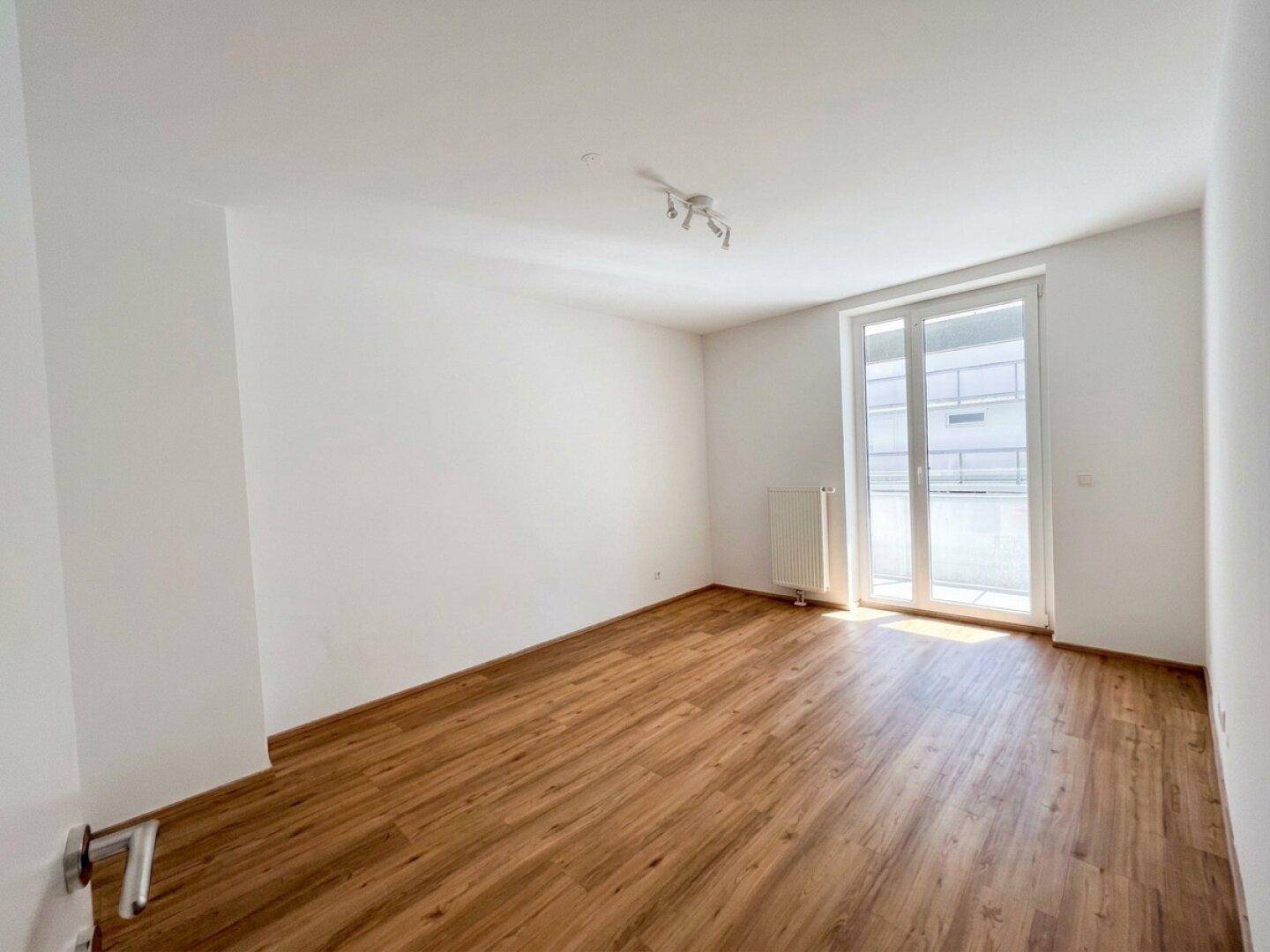 Helles Schlafzimmer mit Holzboden und großem Fenster, das viel Tageslicht hereinlässt.