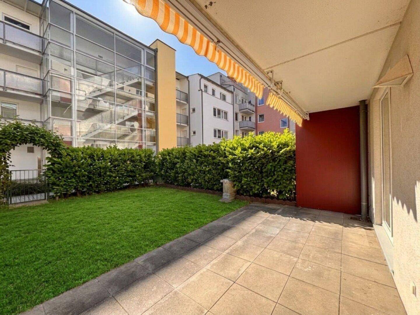 Sonniger Terrassenbereich mit Fliesen und kleiner Rasenfläche, umgeben von grünen Hecken.