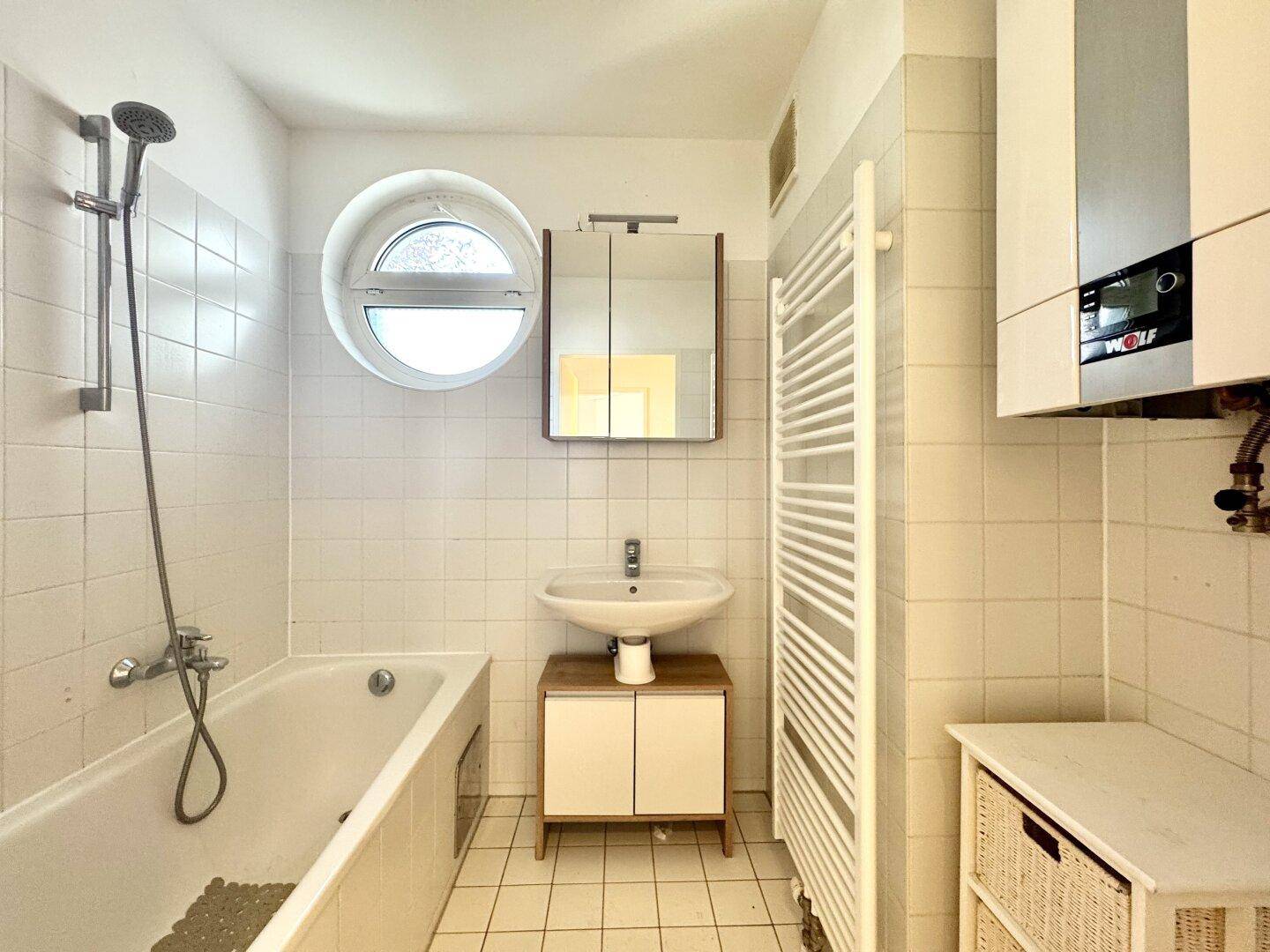 Helles Badezimmer mit Badewanne, runden Fenster, Waschbecken und Handtuchheizkörper.