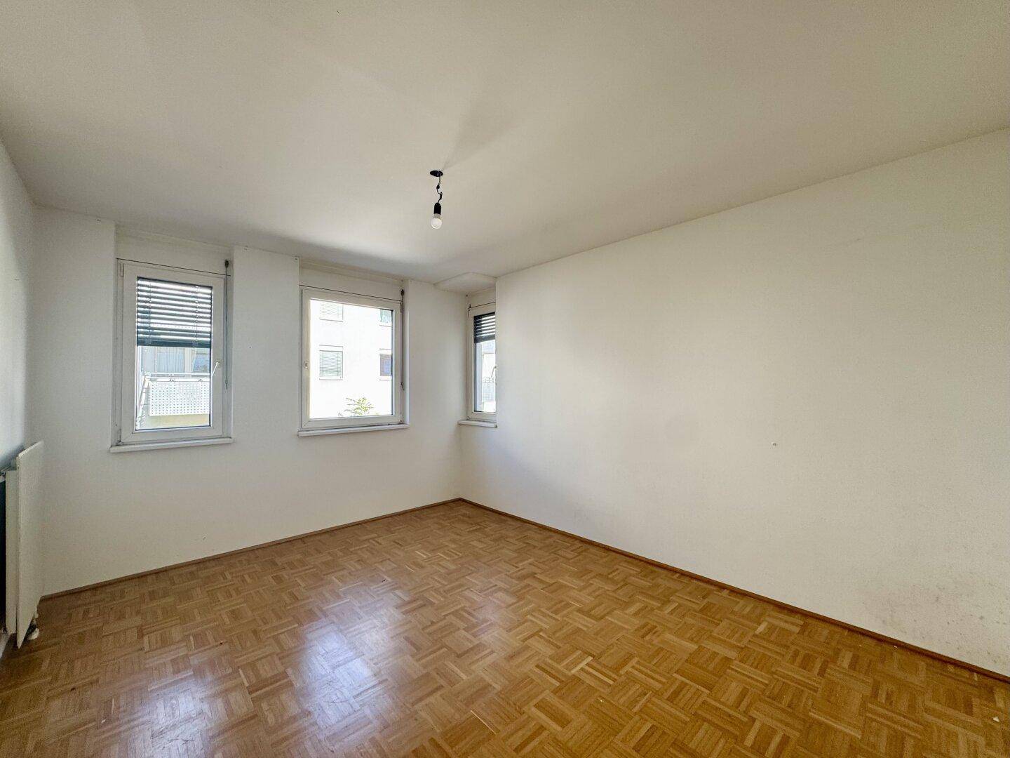 Geräumiges, leeres Schlafzimmer mit Parkettboden und drei Fenstern, ideal für Ruhe und Erholung.