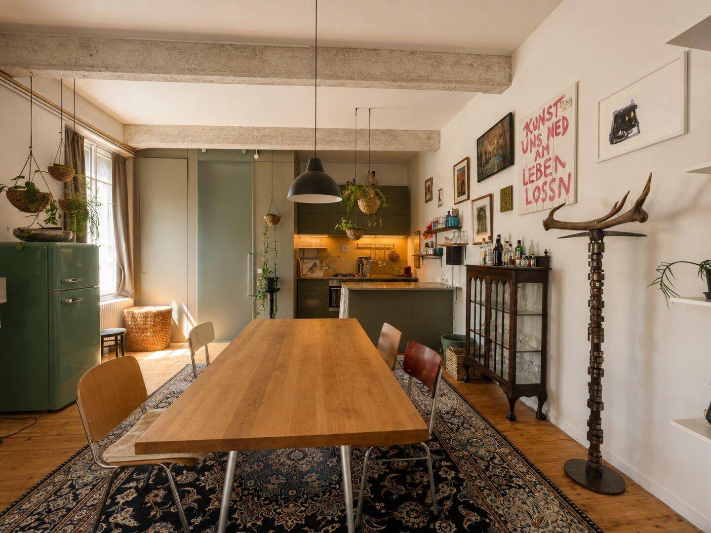 Essbereich mit großem Holztisch und Blick auf die moderne, dunkelgrüne Küche mit integrierten Geräten.