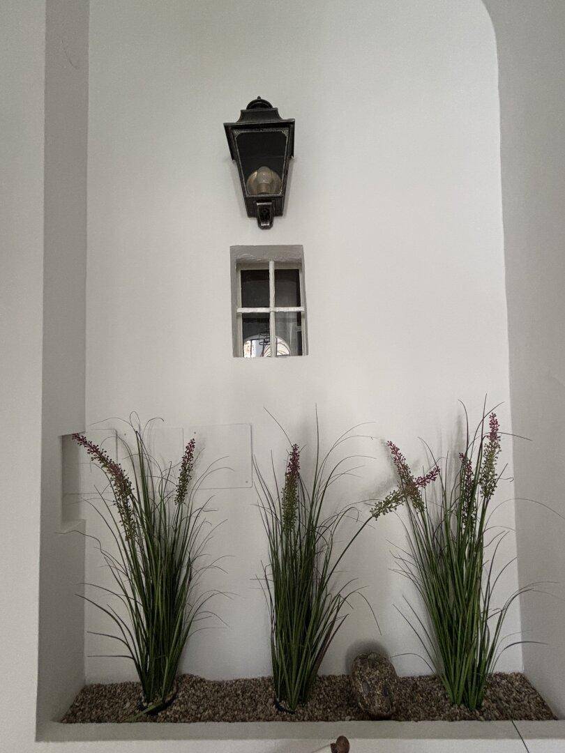 Dekorative Nische mit Kunstpflanzen, einem kleinen Fenster und einer historischen Wandleuchte, schafft Ambiente.