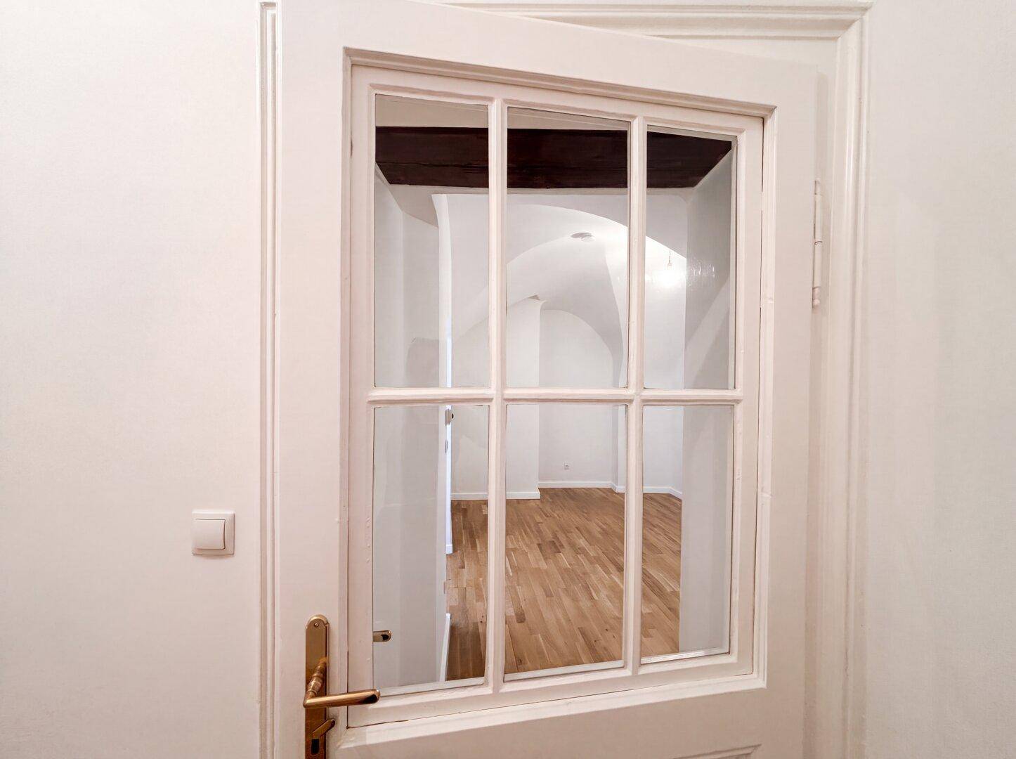 Weiße Tür mit Glaseinsätzen, die den Blick in einen hellen Raum mit Gewölbedecke freigibt.
