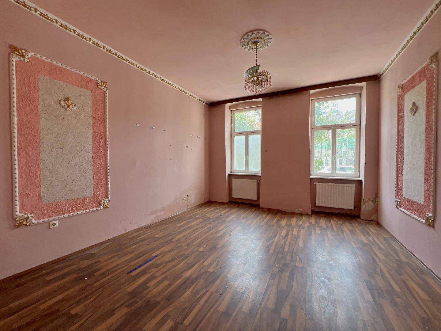 Geräumiges Zimmer mit rosa Wänden, Holzboden, Stuckdecke und zwei großen Fenstern.