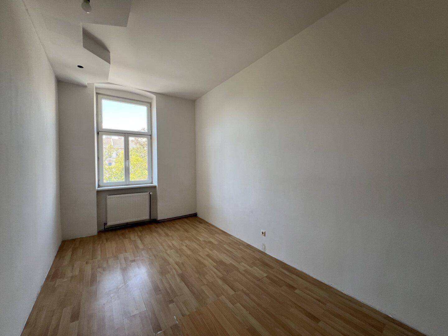 Helles, leeres Zimmer mit Parkettboden und einem großen Fenster, ideal als Schlafzimmer.