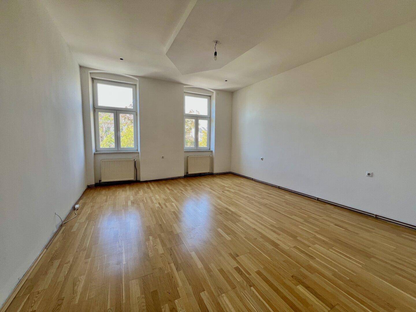 Geräumiges, helles Zimmer mit Parkettboden und zwei Fenstern, ideal als Wohnbereich.