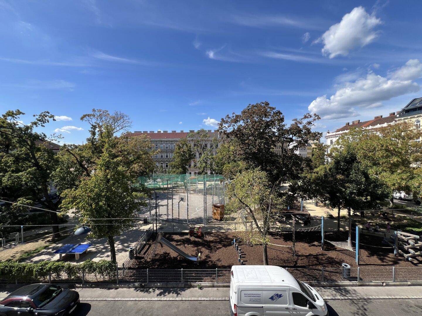 Sonniger Blick auf einen öffentlichen Spielplatz mit Bäumen und umliegenden Gebäuden.