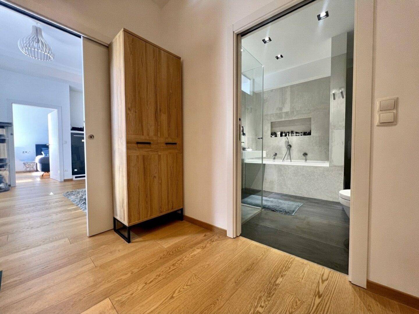 Heller Flur mit Holzboden und Zugang zu einem modernen Badezimmer durch eine Glastür.