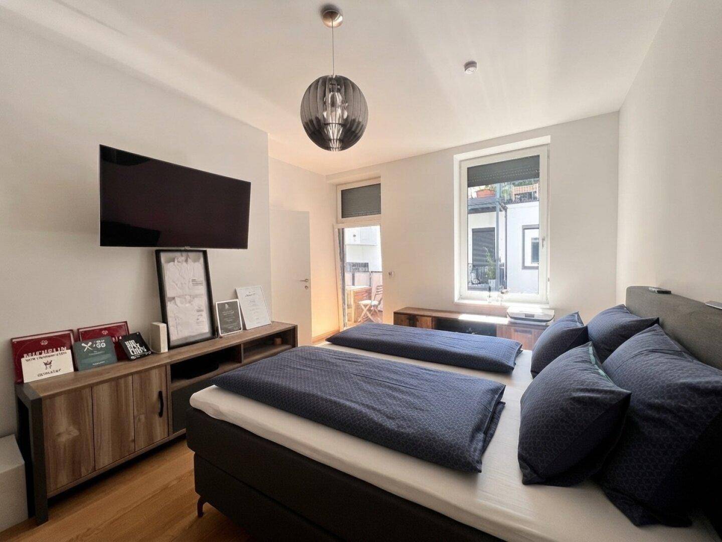 Schlafzimmer mit Doppelbett, Wandfernseher und direktem Zugang zu einem sonnigen Balkon.