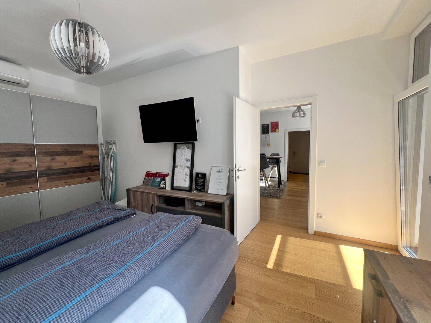 Schlafzimmer mit Doppelbett, Wandfernseher und Zugang zu einem weiteren Wohnbereich.