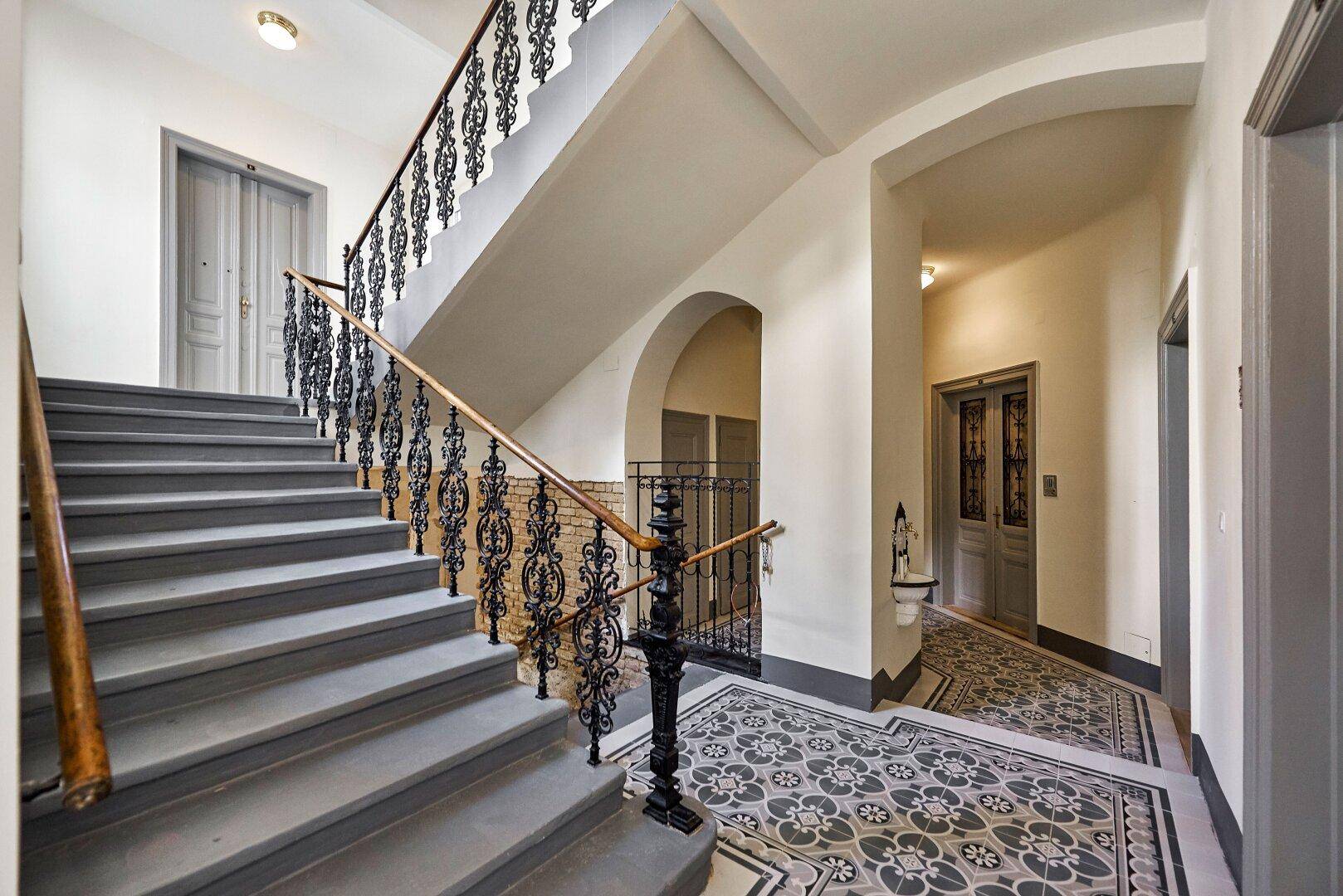 Elegantes Treppenhaus mit schmiedeeisernem Geländer, gemusterten Fliesen und hohen Decken.