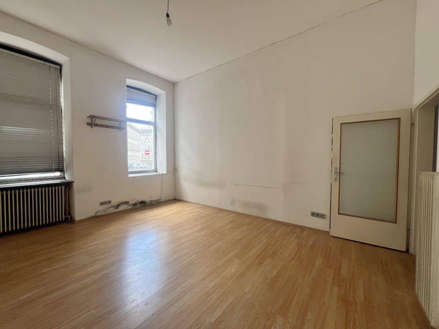 Helles Zimmer mit Parkettboden, zwei Fenstern und Heizkörper unter den Fenstern.