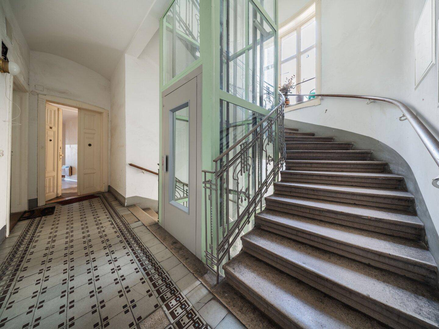 Historisches Treppenhaus mit modernem Aufzug und gemustertem Fliesenboden im Eingangsbereich.