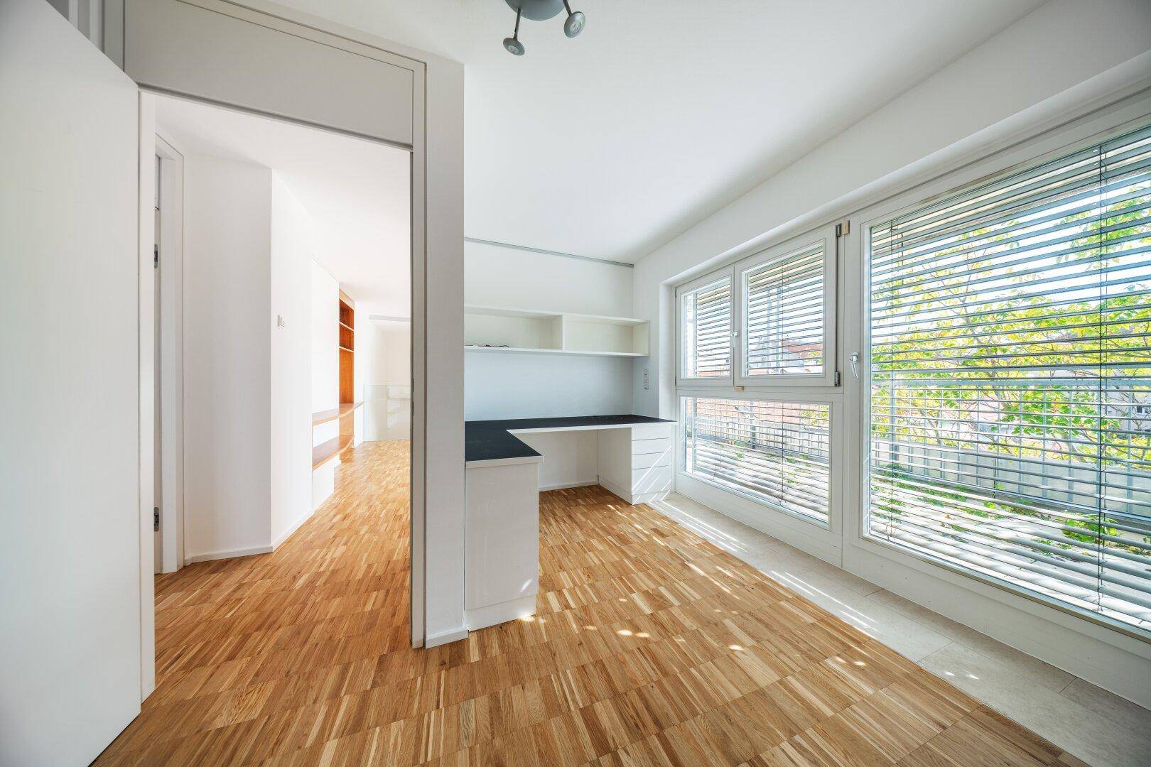 Helles Arbeitszimmer mit Parkettboden, großen Fenstern und integriertem Schreibtisch.