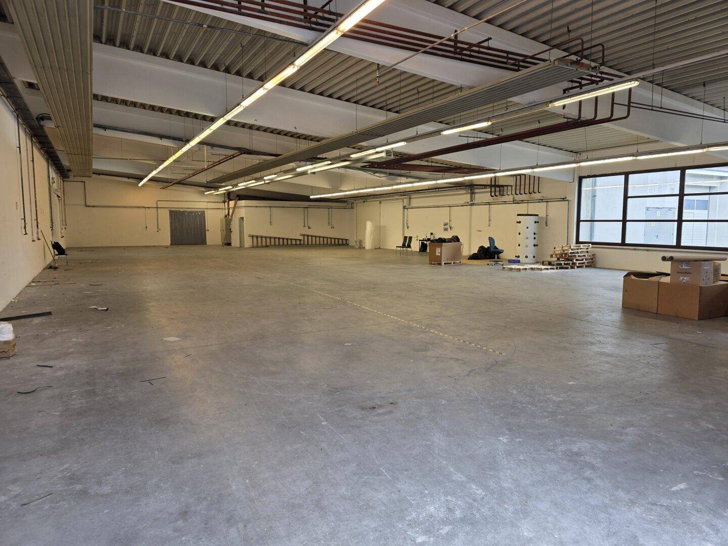 Große, helle Lagerhalle mit Betonboden, hohen Decken und Fenstern, die viel Tageslicht hereinlassen.