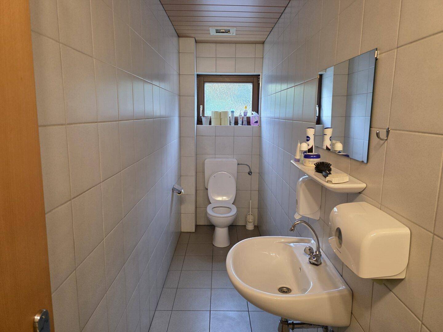 Toilettenbereich mit WC, Waschbecken und Spiegel, komplett gefliest und mit Fenster ausgestattet.