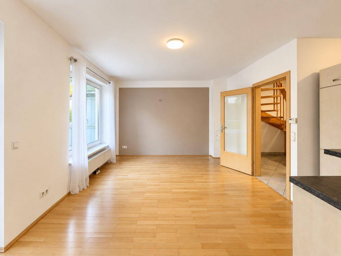 Heller Wohnbereich mit Holzboden, großem Fenster und offenem Zugang zur Treppe und Küche.