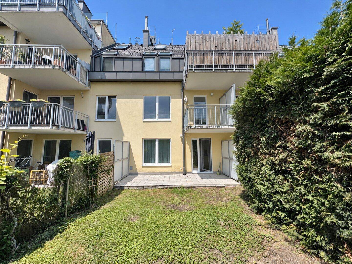 Hinteransicht des Gebäudes mit Gartenbereich und unmöblierter Terrasse unter blauem Himmel.