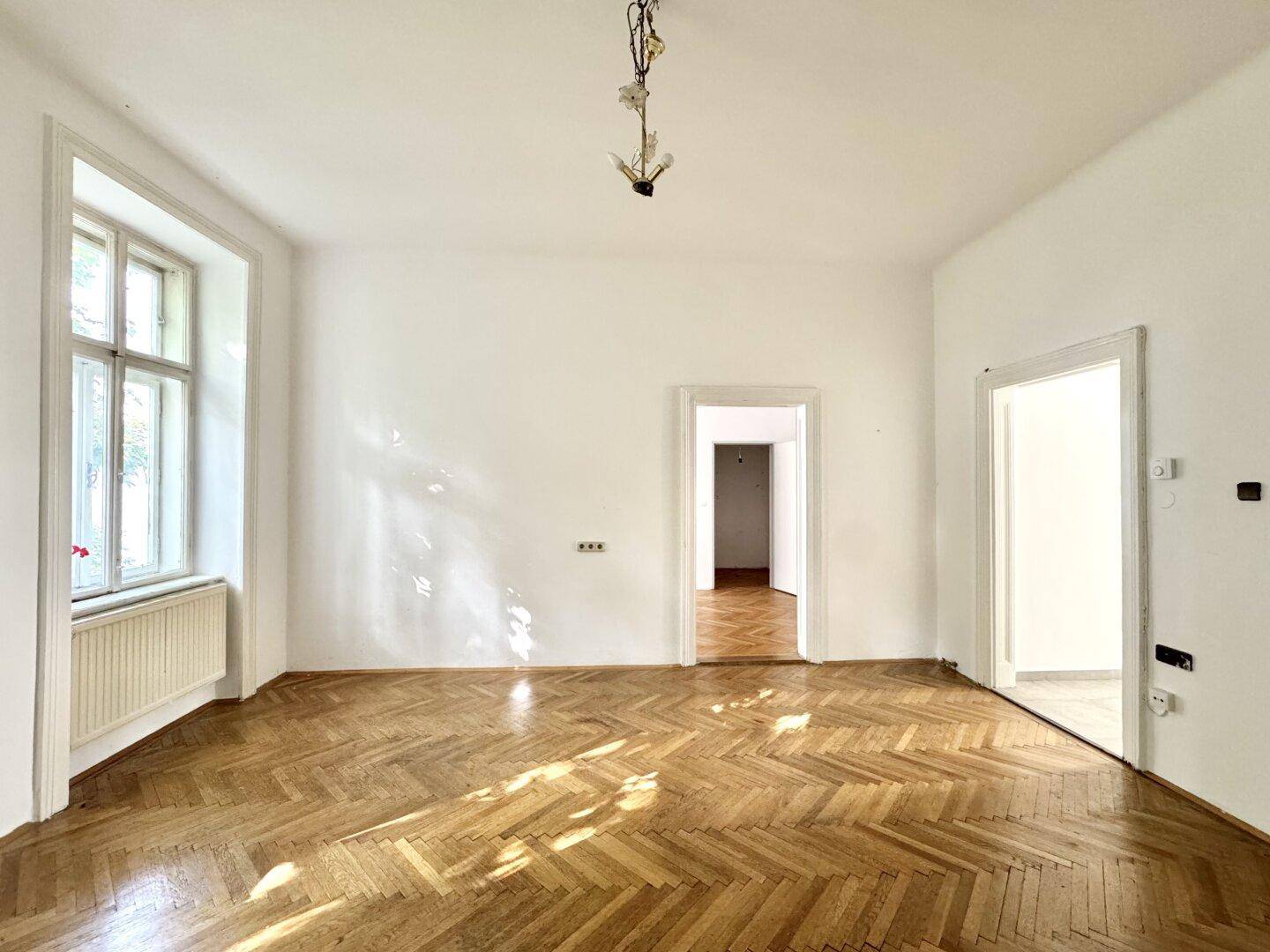 Heller Raum mit Fischgrätparkett und hohen Decken, der auf weitere Zimmer blicken lässt.