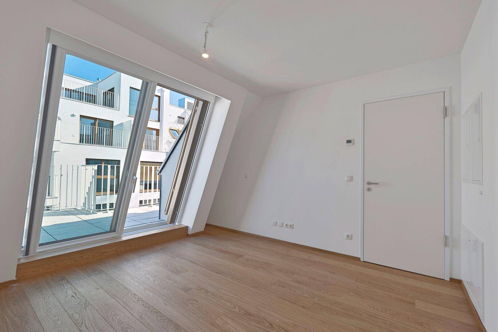 Geräumiger, heller Raum mit Holzboden und großem Fenster, das Zugang zur Außenterrasse bietet.
