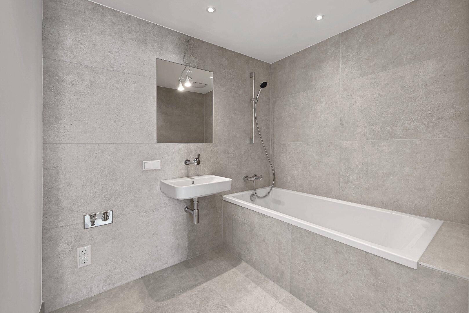 Zeitgemäßes Badezimmer mit Badewanne, kleinem Waschbecken und grauen Wandfliesen.