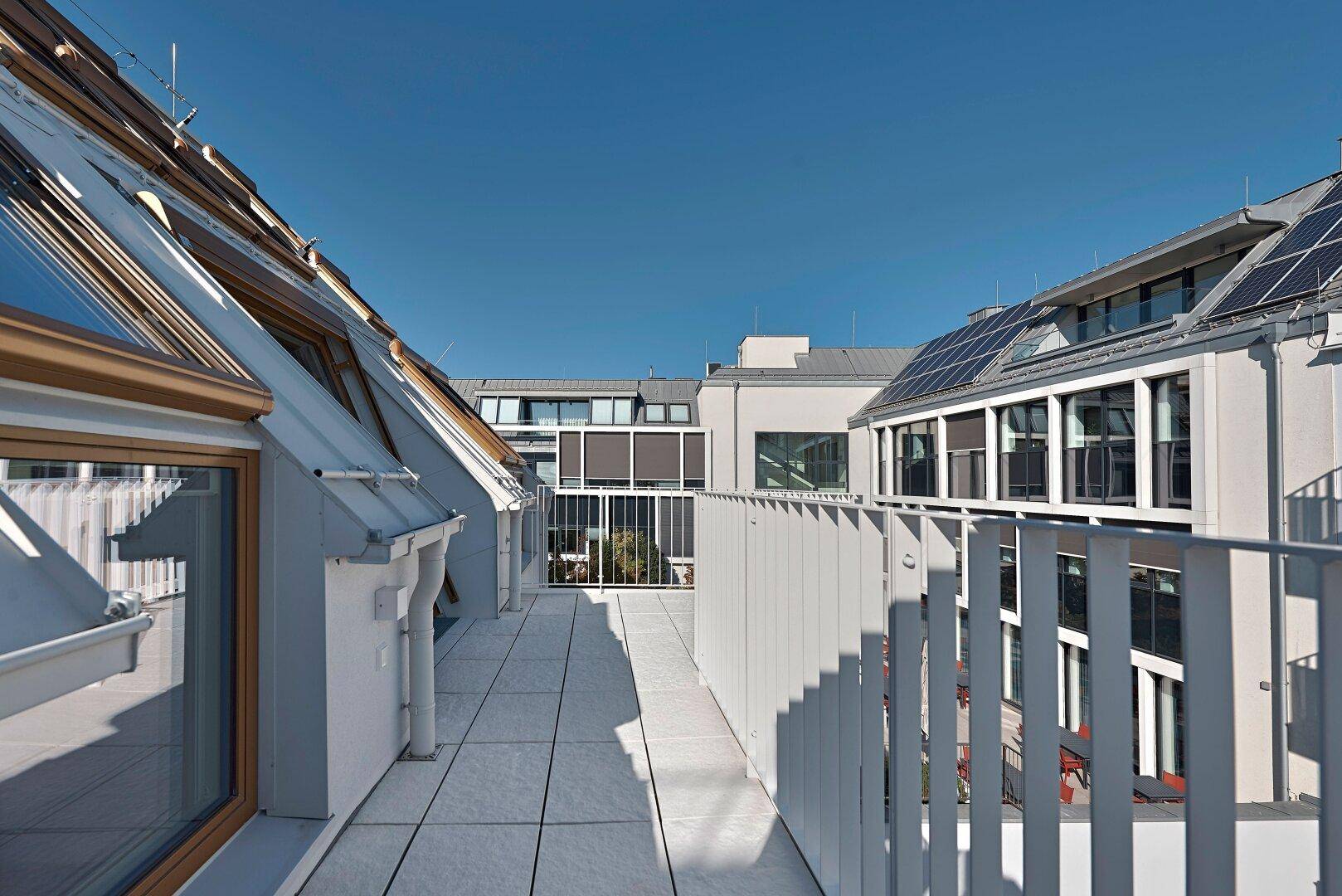 Lange Dachterrasse mit Fliesenboden, umgeben von modernen Gebäuden und Solaranlagen.