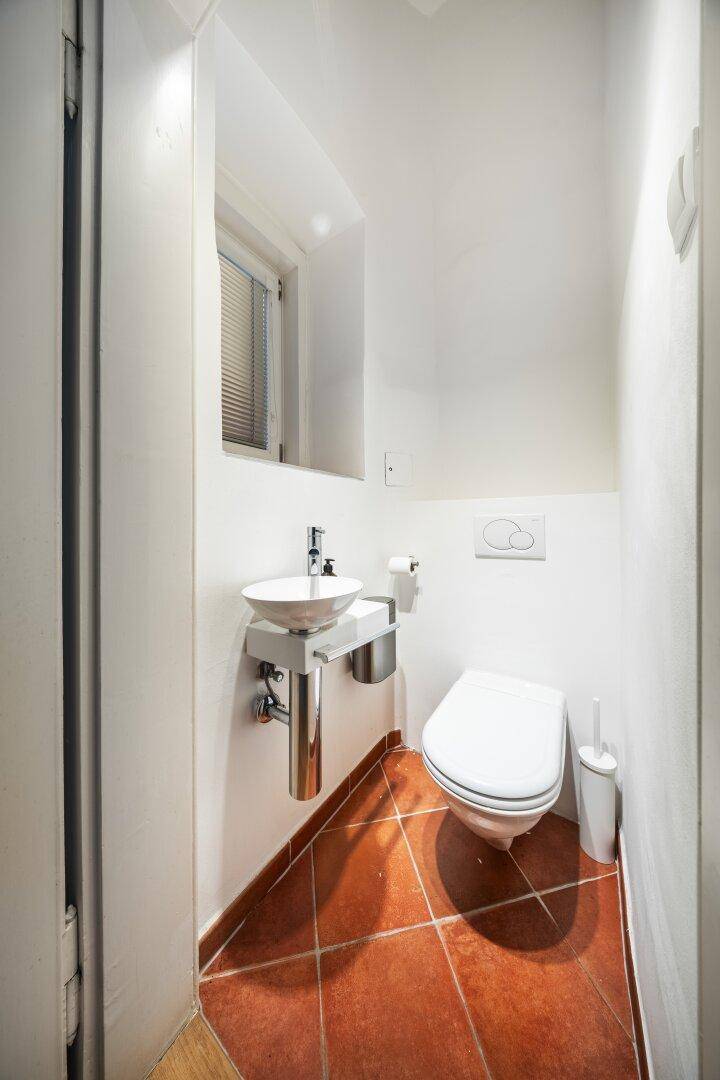 Kleines Gäste-WC mit Terrakotta-Fliesenboden, wandhängendem WC und kleinem Waschbecken.